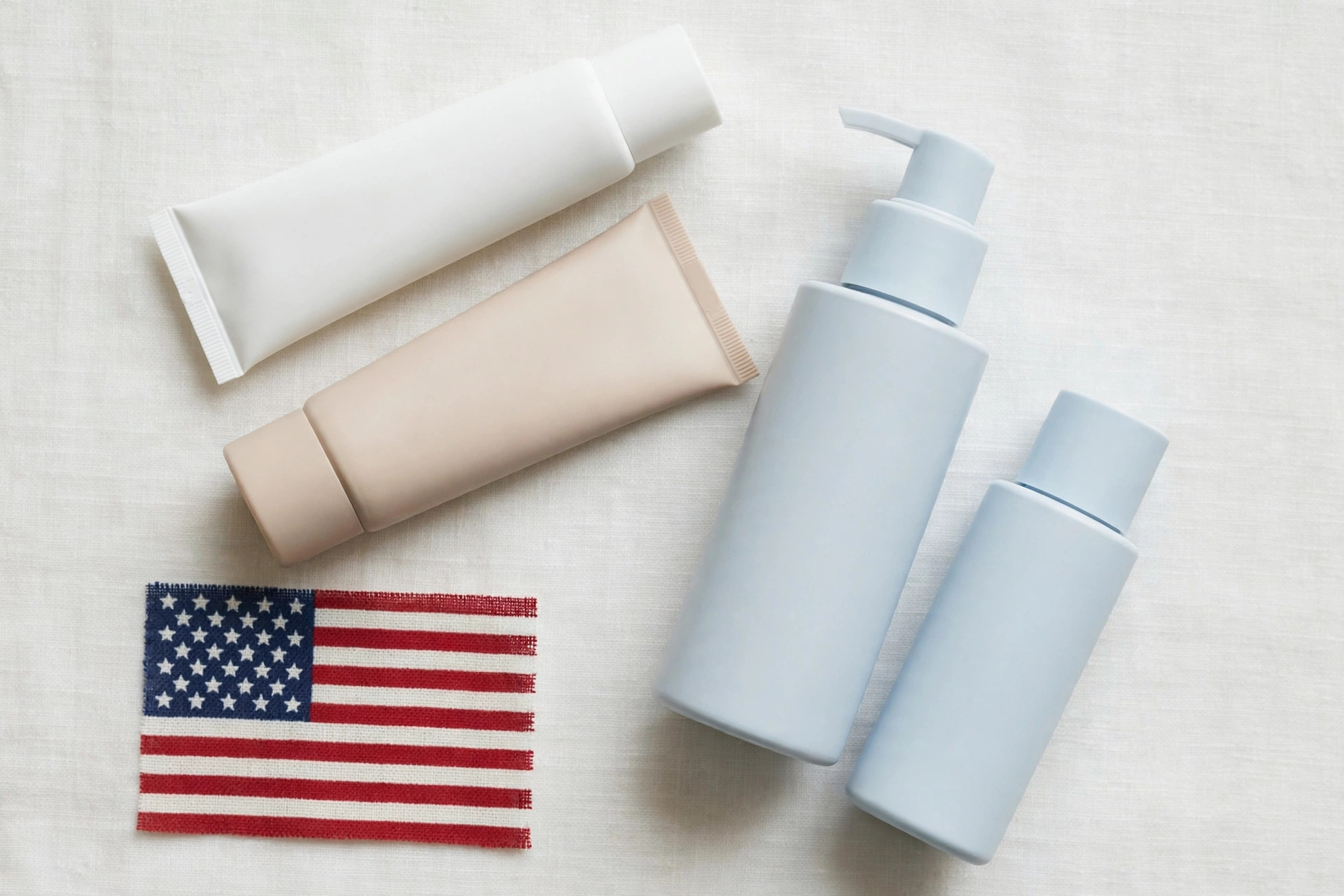 Unlabeled sunscreen tubes and bottles arranged on a light background beside a small American flag fabric swatch